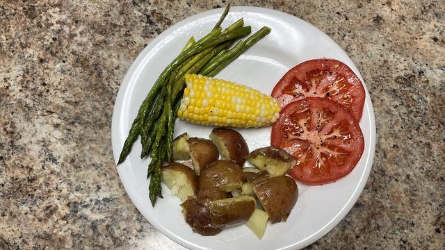 White dinner plate with roasted asparagus, corn on the cob, boiled red skinned potatoes, and sliced tomatoes.