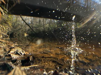 Low-angle view of a shallow stream beneath a footbridge, sunlight catching water droplets mid-splash; fallen leaves and wet stones in the foreground with bare trees and blue sky blurred in the background.