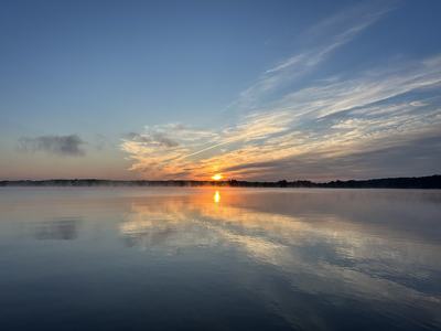Tranquil sunrise over a misty lake: a golden sun peeks above a dark treeline, streaked clouds and warm light reflected on calm, glassy water.