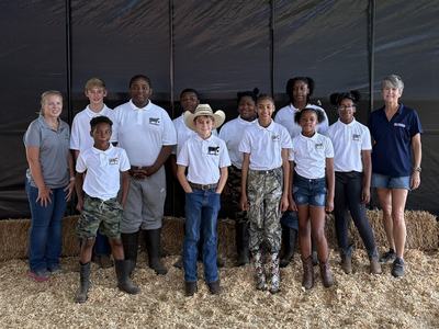 A large group of people—mostly youth with two adults—stand posing for a photo on a ground covered in wood shavings or mulch, in front of a large black backdrop and hay bales.