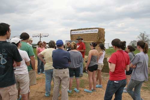 People listening to a guide by stacked hay bales with sign "AGRICULTURE EDUCATION CONCERN"