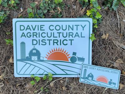 A rectangular sign reading “Davie County Agricultural District." The sign features a stylized farm scene with a barn, silo, tractor, and a rising sun over fields. A smaller matching sign in front of it