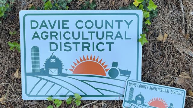 A rectangular sign reading “Davie County Agricultural District." The sign features a stylized farm scene with a barn, silo, tractor, and a rising sun over fields. A smaller matching sign in front of it