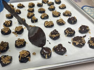 spoon dipped in chocolate over sheet pan of coconut crunch stuffed prunes