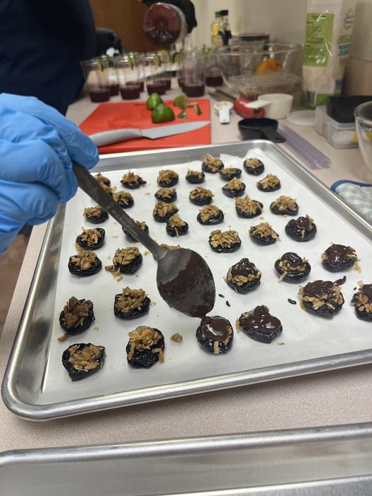 spoon dipped in chocolate over sheet pan of coconut crunch stuffed prunes
