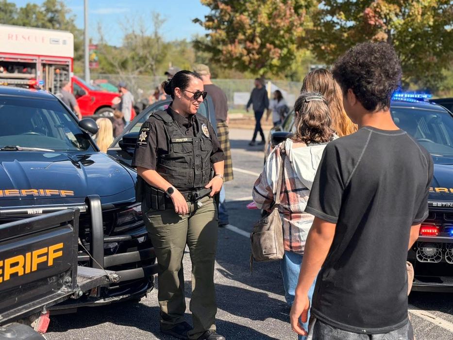 A smiling sheriff's deputy speaks with community members next to a patrol vehicle.