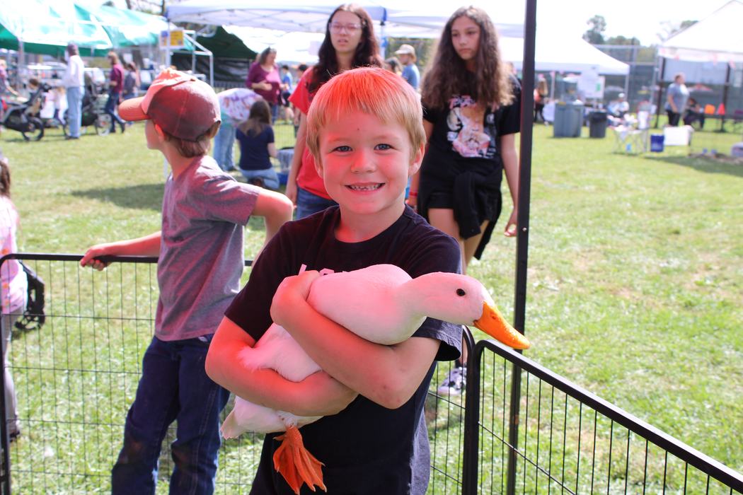 Young boy smiling while holding a large white duck at an outdoor fair.