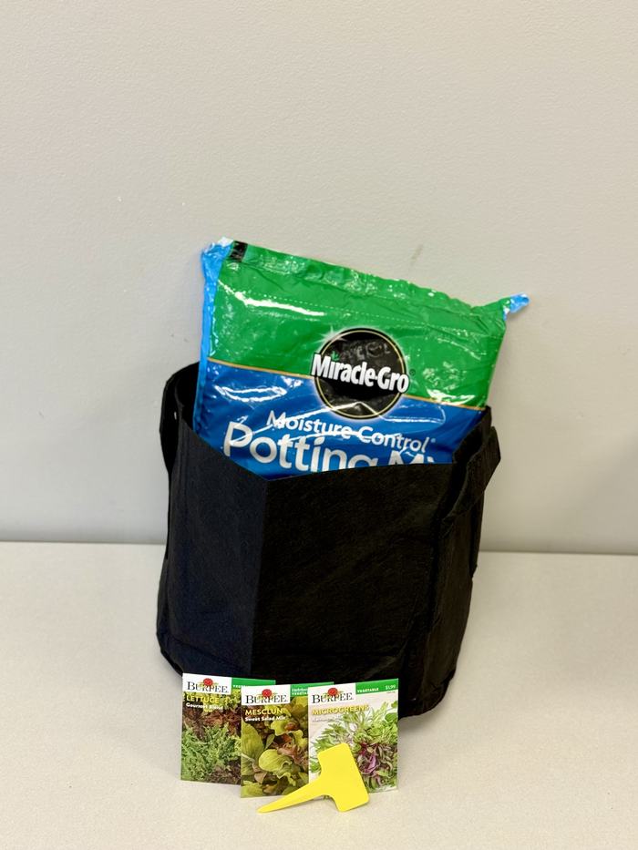 Black 5-gallon grow bag filled with potting mix and a bag of potting soil, displayed with vegetable seed packets and a small planting tool provided to participants in the Seed to Supper gardening program.