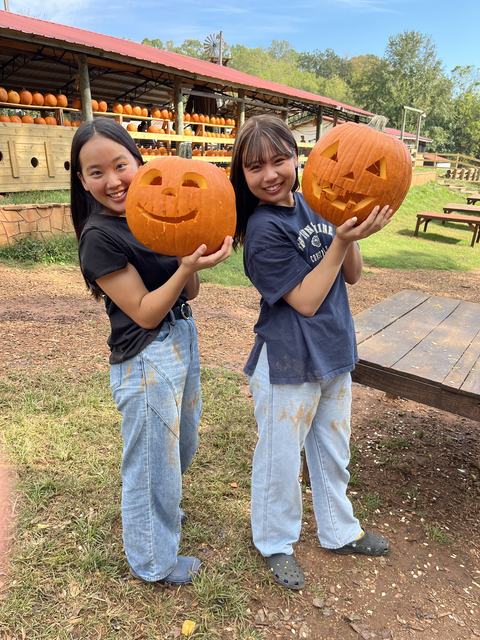 Two Japanese high school students stand next to each other and hold up large jack-o-lanterns that they carved for Halloween.