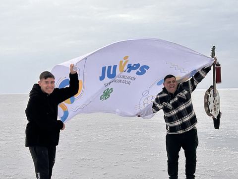 A man and a teenage boy stand in the salt flats in Argentina holding a JUVIPS Argentina 4-H flag between them. The man is holding a musical instrument made of recycled materials and a water bottle in his other hand.
