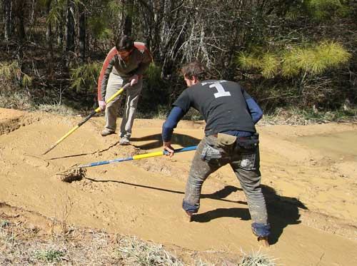 Two people leveling a dirt path with rakes; back of one shirt shows the number "1"