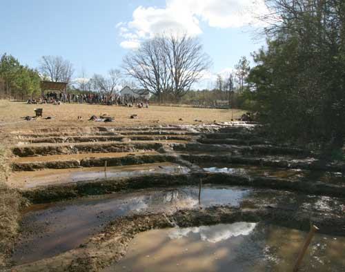 Tiered muddy pools with shallow water in foreground; crowd by pavilion and trees in background