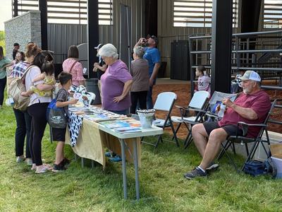 A group of people, including children holding cow-themed masks, stand around an outdoor information table covered in a cow-print cloth. An older man sits in a folding chair nearby under the shade of a large pavilion.