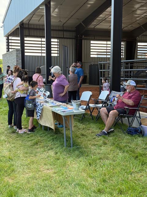 A group of people, including children holding cow-themed masks, stand around an outdoor information table covered in a cow-print cloth. An older man sits in a folding chair nearby under the shade of a large pavilion.