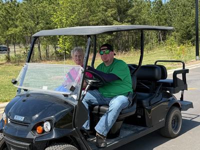 A man in a green t-shirt and sunglasses drives a black E-Z-GO golf cart on a paved path, with a woman in a pink shirt riding as a passenger. A line of pine trees and a clear blue sky are in the background.