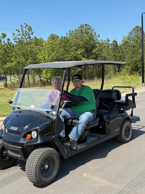 A man in a green t-shirt and sunglasses drives a black E-Z-GO golf cart on a paved path, with a woman in a pink shirt riding as a passenger. A line of pine trees and a clear blue sky are in the background.