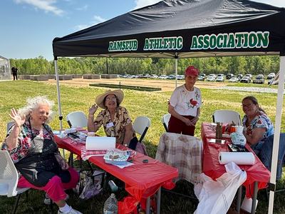 Four women sit and stand under a black tent on a sunny day. They are positioned behind tables covered in red cloths with various supplies, and several of them are waving at the camera.