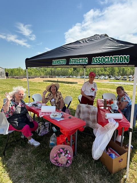 Four women sit and stand under a black tent on a sunny day. They are positioned behind tables covered in red cloths with various supplies, and several of them are waving at the camera.