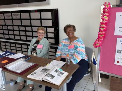 Two women sit smiling at a registration desk inside the Glen Ray McNeill Education & Resource Center. Papers and an open book are on the table in front of them, and a pink display board decorated with a floral lei stands to the right.