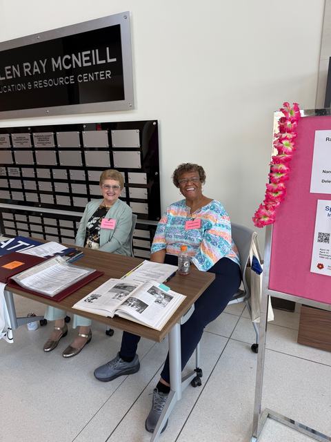 Two women sit smiling at a registration desk inside the Allen Ray McNeill Education & Resource Center. Papers and an open book are on the table in front of them, and a pink display board decorated with a floral lei stands to the right.