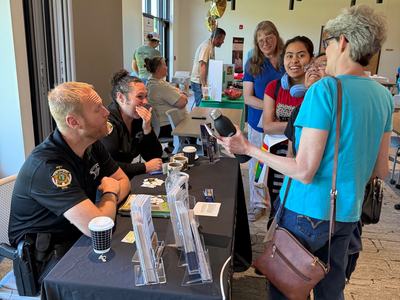 Two law enforcement officers sit behind a black draped table, talking and laughing with a group of visitors inside a modern building with a unique geometric ceiling. Pamphlets and coffee cups are on the table.