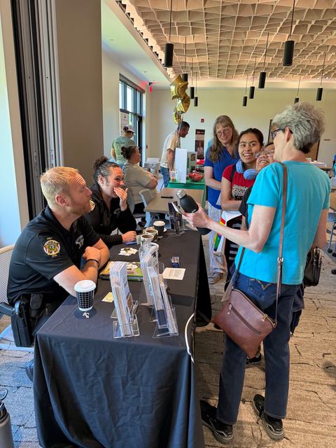 Two law enforcement officers sit behind a black draped table, talking and laughing with a group of visitors inside a modern building with a unique geometric ceiling. Pamphlets and coffee cups are on the table.