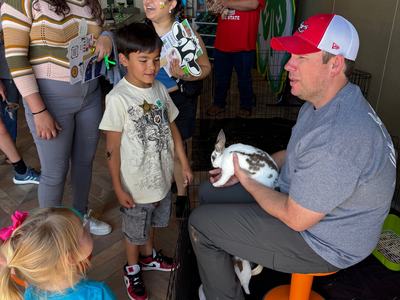 A man sits on a stool inside a wire pen, holding a spotted rabbit for a group of children and adults to see. A large green 4-H clover backdrop is visible in the corner of the room.