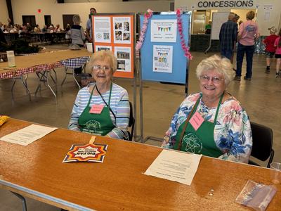 Two women wearing green aprons and name tags sit smiling behind a wooden registration table at an event. Behind them are informational displays, and the large indoor venue features a concessions area and people sitting at long tables in the background.