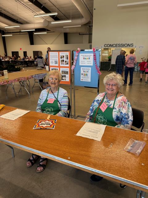 Two women wearing green aprons and name tags sit smiling behind a wooden registration table at an event. Behind them are informational displays, and the large indoor venue features a concessions area and people sitting at long tables in the background.