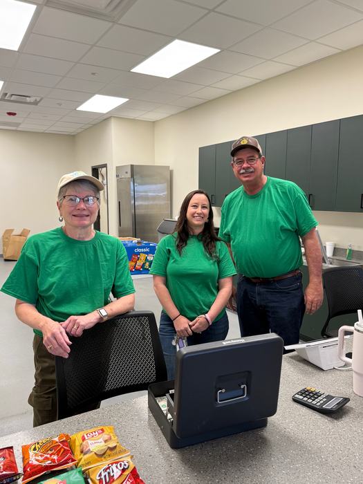 Three people wearing matching green t-shirts stand behind a counter in a bright room, likely a concessions area. An open cash box, a calculator, and various bags of snacks are visible on the counter in the foreground.