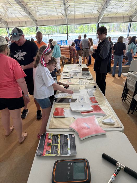 young people stand at a long table in a large, open-sided pavilion, interacting with a puzzle of a cow's body that shows different cuts of meat. Other people and a livestock pen are visible in the background.