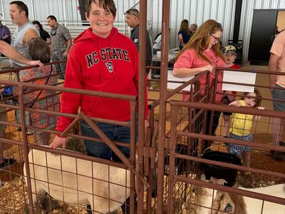 A boy in a red NC State hoodie smiles while standing in a livestock pen and petting a sheep. A goat stands in an adjacent pen, and several people are visible in the background of the indoor arena.
