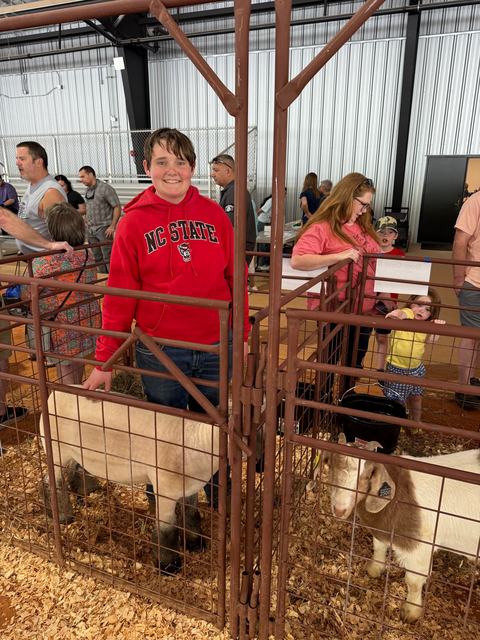A boy in a red NC State hoodie smiles while standing in a livestock pen and petting a sheep. A goat stands in an adjacent pen, and several people are visible in the background of the indoor arena.
