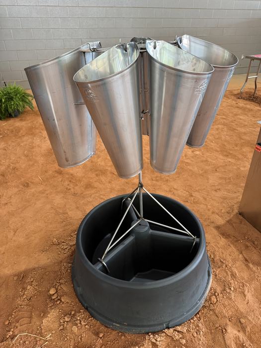 A specialized poultry processing station featuring a circular arrangement of stainless steel cones suspended over a large black plastic collection tub on a dirt floor.