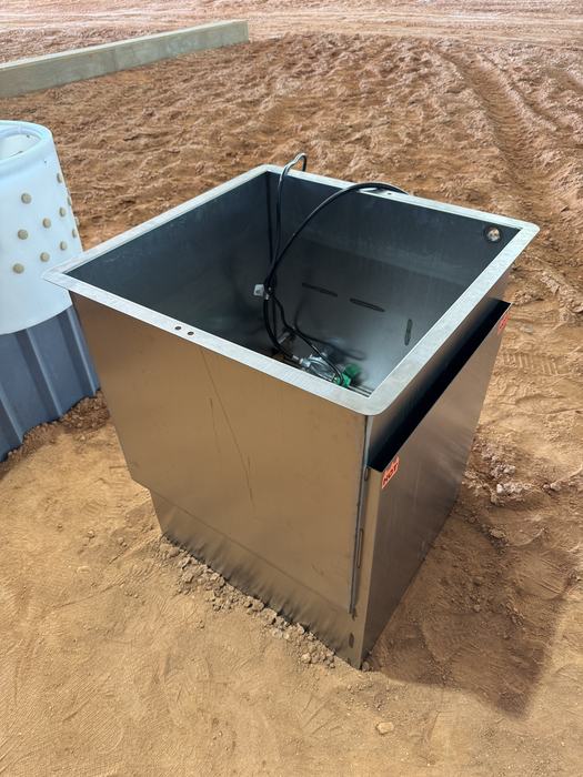 A large, square stainless steel scalding tank sits on a red dirt floor, with internal heating elements and wiring visible inside the basin.