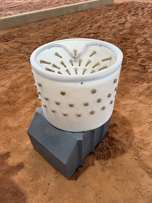 A motorized poultry plucker featuring a white plastic tub lined with small rubber fingers, mounted on a grey base over a red dirt floor.