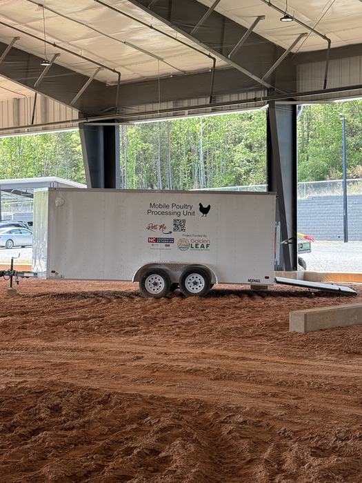 A white tandem-axle trailer labeled "Mobile Poultry Processing Unit" parked on the dirt floor of an open-sided indoor arena. The trailer features logos for the NC Cooperative Extension and the Golden LEAF Foundation, along with a "Rent Me" graphic