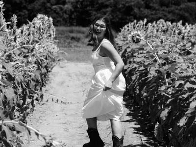 Black-and-white photo of a smiling woman in a white sundress and cowboy boots skipping down a dirt path between tall sunflower rows, hair flowing and laughing as she looks back mid-turn.
