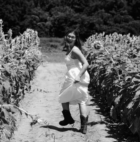 Black-and-white photo of a smiling woman in a white sundress and cowboy boots skipping down a dirt path between tall sunflower rows, hair flowing and laughing as she looks back mid-turn.