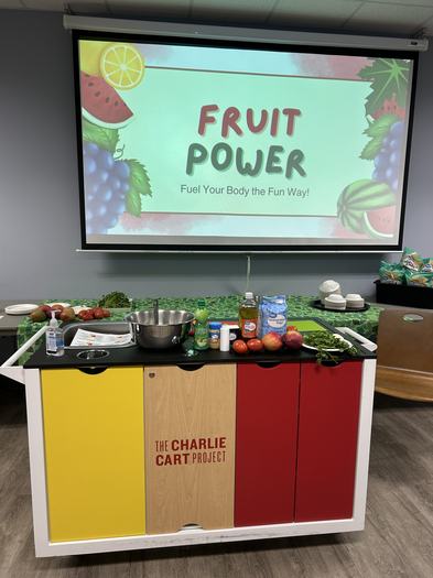 The Charlie Cart set up with ingredients for a cooking demo in front of a presentation screen that says "Fruit Power: Fuel your body the fun way!"