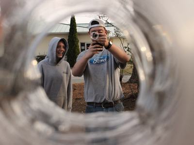 View through a circular tube shows two teens outdoors; one in a Ford T‑shirt and backward cap filming toward the viewer with a camcorder, while a smiling friend in a gray hoodie stands beside him.