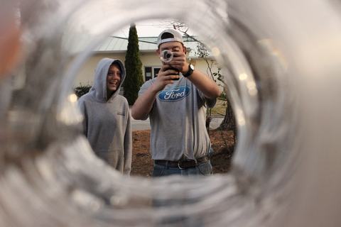View through a circular tube shows two teens outdoors; one in a Ford T‑shirt and backward cap filming toward the viewer with a camcorder, while a smiling friend in a gray hoodie stands beside him.