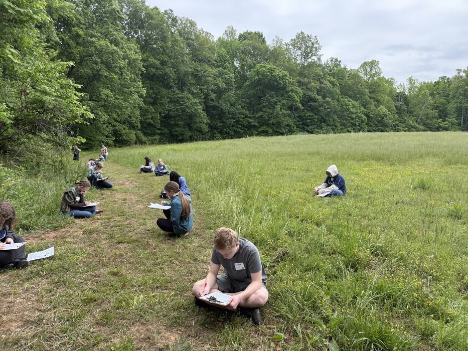 Youth are seated in a grassy field with clipboards