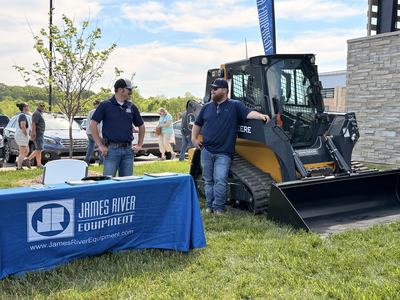 Two men stand outside on a grassy area next to a blue table with a James River Equipment logo; one man leans against a yellow and black John Deere skid steer loader while talking to the other.