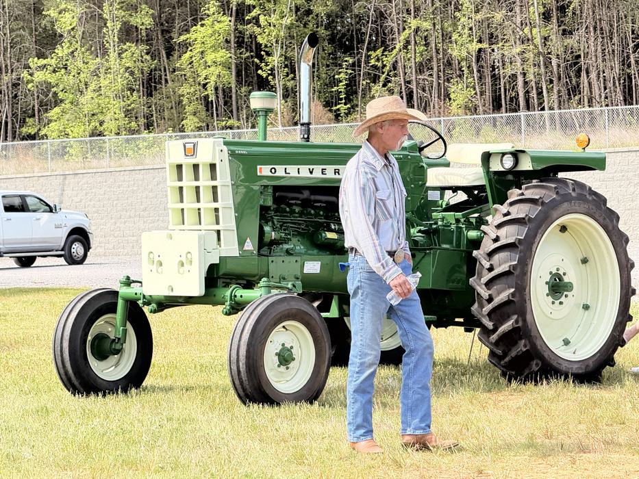 A man in a straw hat, button-down shirt, and jeans walks past a vintage green and white Oliver tractor parked on a grassy field. Woods and a white pickup truck are visible in the background.
