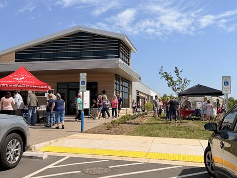 A crowd of people walks along a sidewalk and grassy area in front of a modern agricultural center building during an outdoor festival with red and black event tents.
