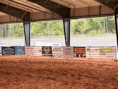 A row of colorful sponsor banners hangs along the fence of an indoor dirt arena, featuring local businesses and organizations like Tarheel Metal Structures and the Randolph County Agricultural Center.