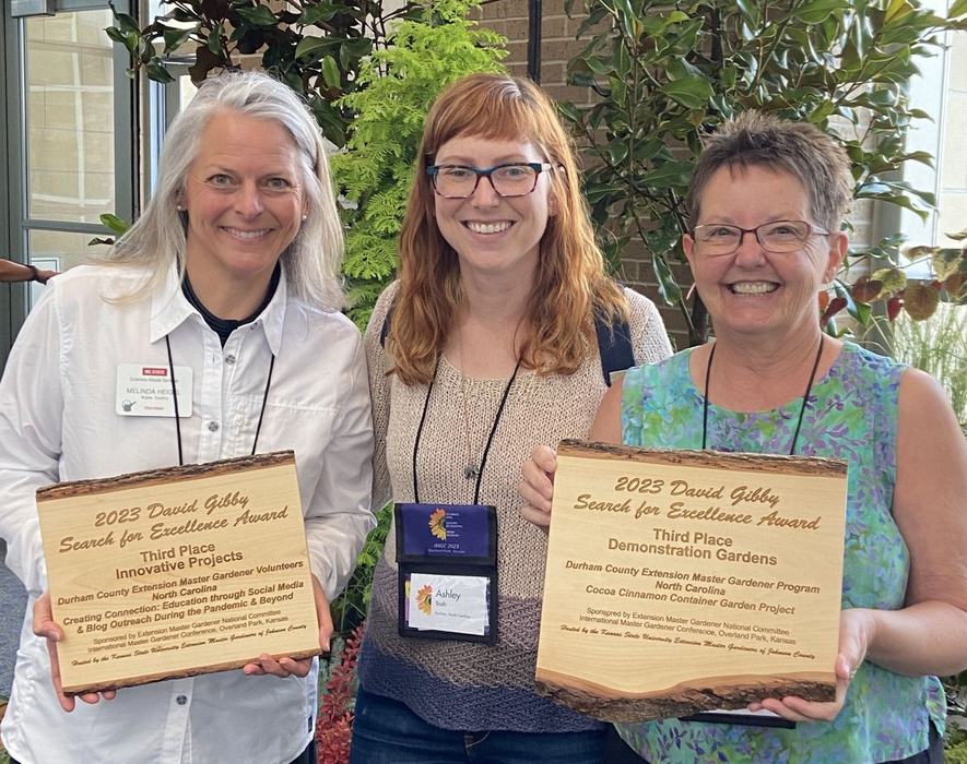 Three women display wooden plaques bearing the inscription "2023 David Gibby Search for Excellence Award."