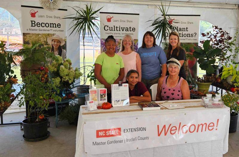 Six women staff a gardening information booth surrounded by plants and Extension Master Gardener volunteer banners.