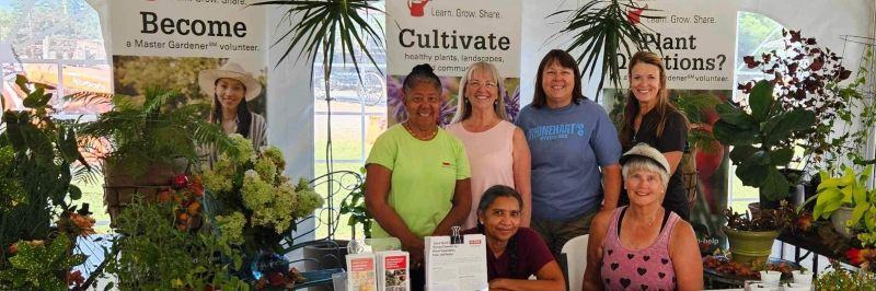 Six women staff a gardening information booth surrounded by plants and Extension Master Gardener banners.